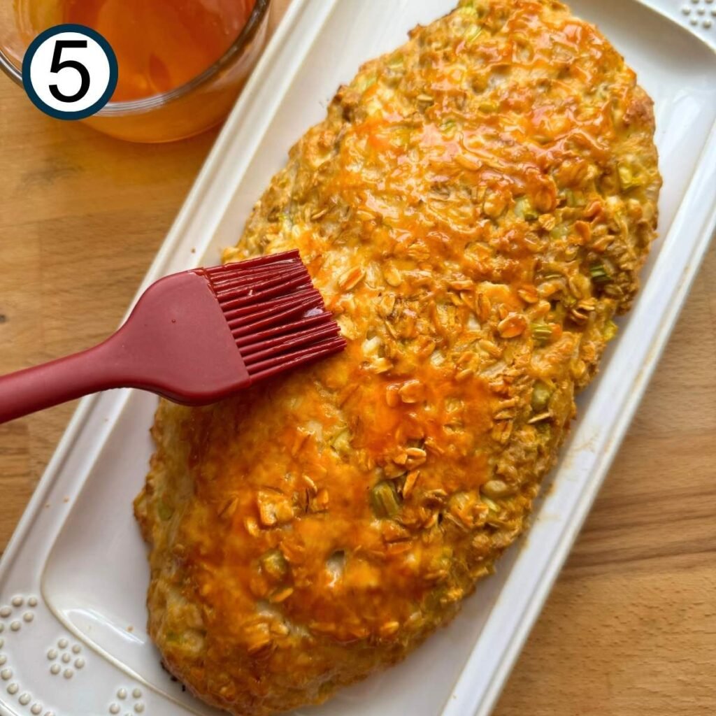 Baked buffalo chicken meatloaf on a platter with a brush adding more buffalo sauce on top.