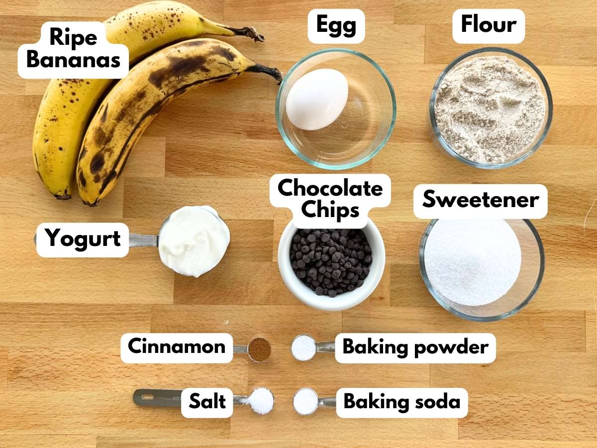 Two ripe bananas, egg, flour, Greek yogurt, mini chocolate chips, sugar and stevia blend, cinnamon, baking powder, baking soda, and salt on a cutting board.