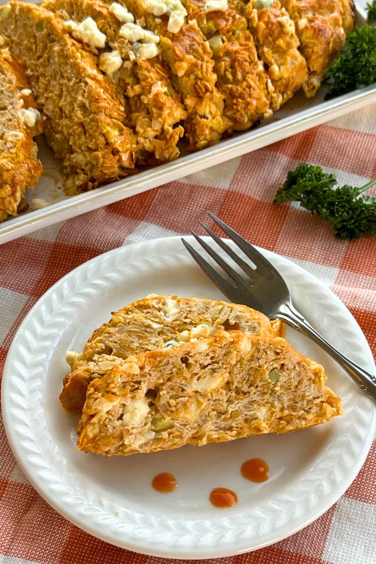 Two slices of buffalo chicken meatloaf on a plate with fork with a platter with more of the loaf behind it.