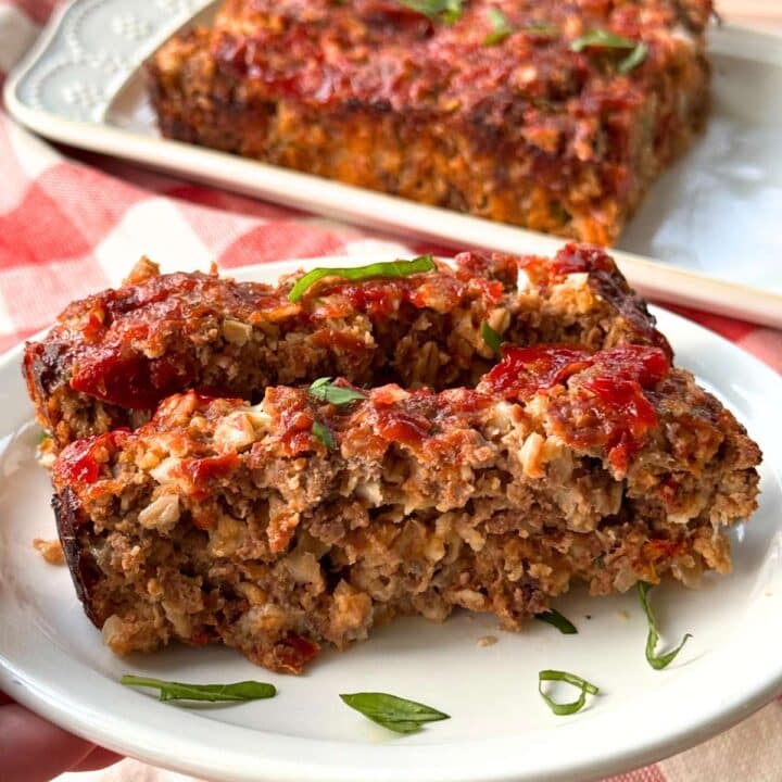 Two slices of gourmet meatloaf with sundried tomatoes, mozzarella and Parmesan cheese, and oatmeal on a white plate with chopped basil.