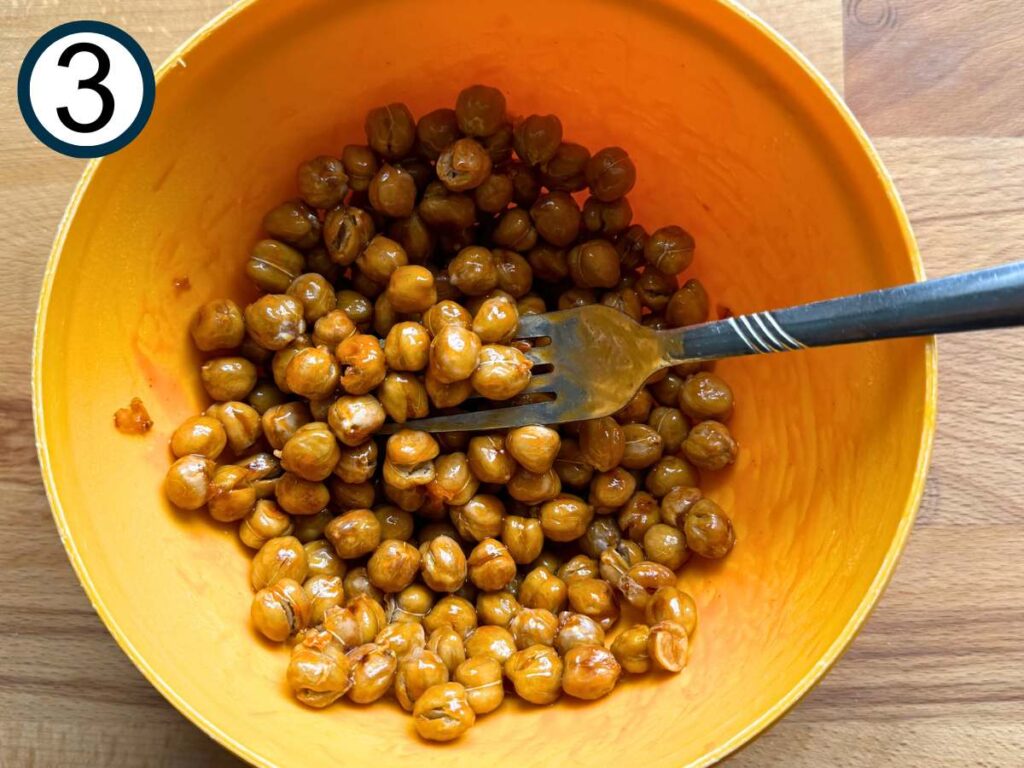 Cooked chickpeas tossed with buffalo sauce in a orange bowl with a fork.