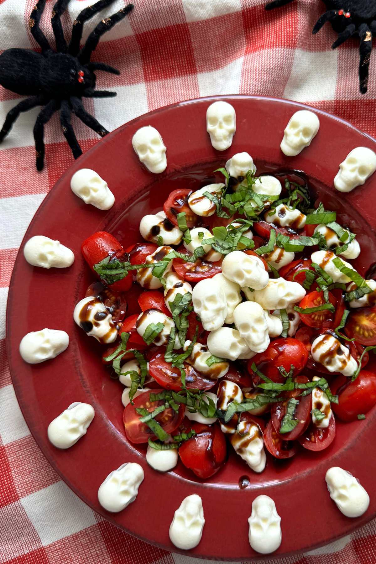 Large red bowl with Halloween caprese salad with mozzarella skulls around the flat edge and piled in the center and with decorative spiders in the background.