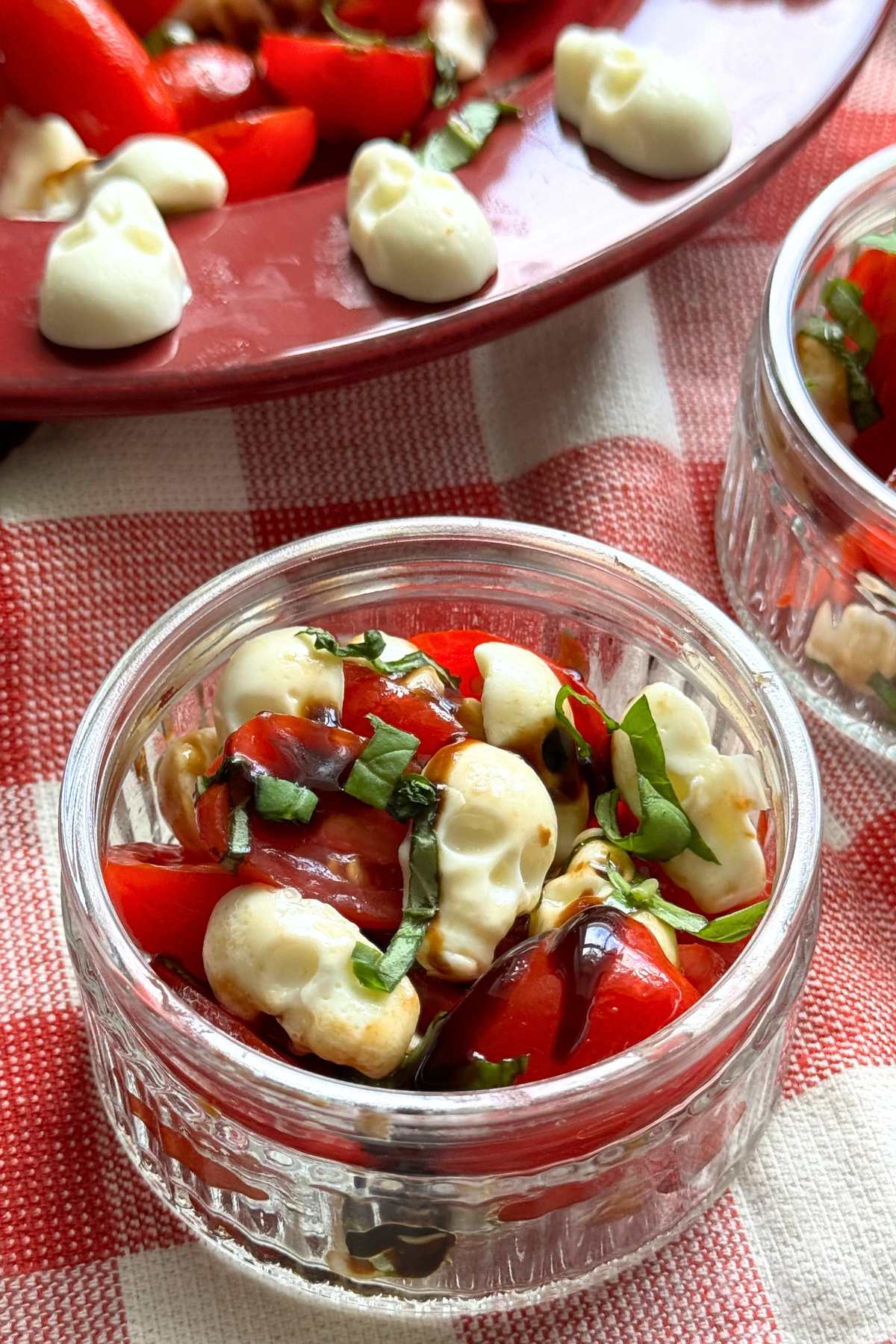 Small glass bowl with a serving of Halloween caprese salad with spooky mozzarella skulls with bowl of full salad behind it.