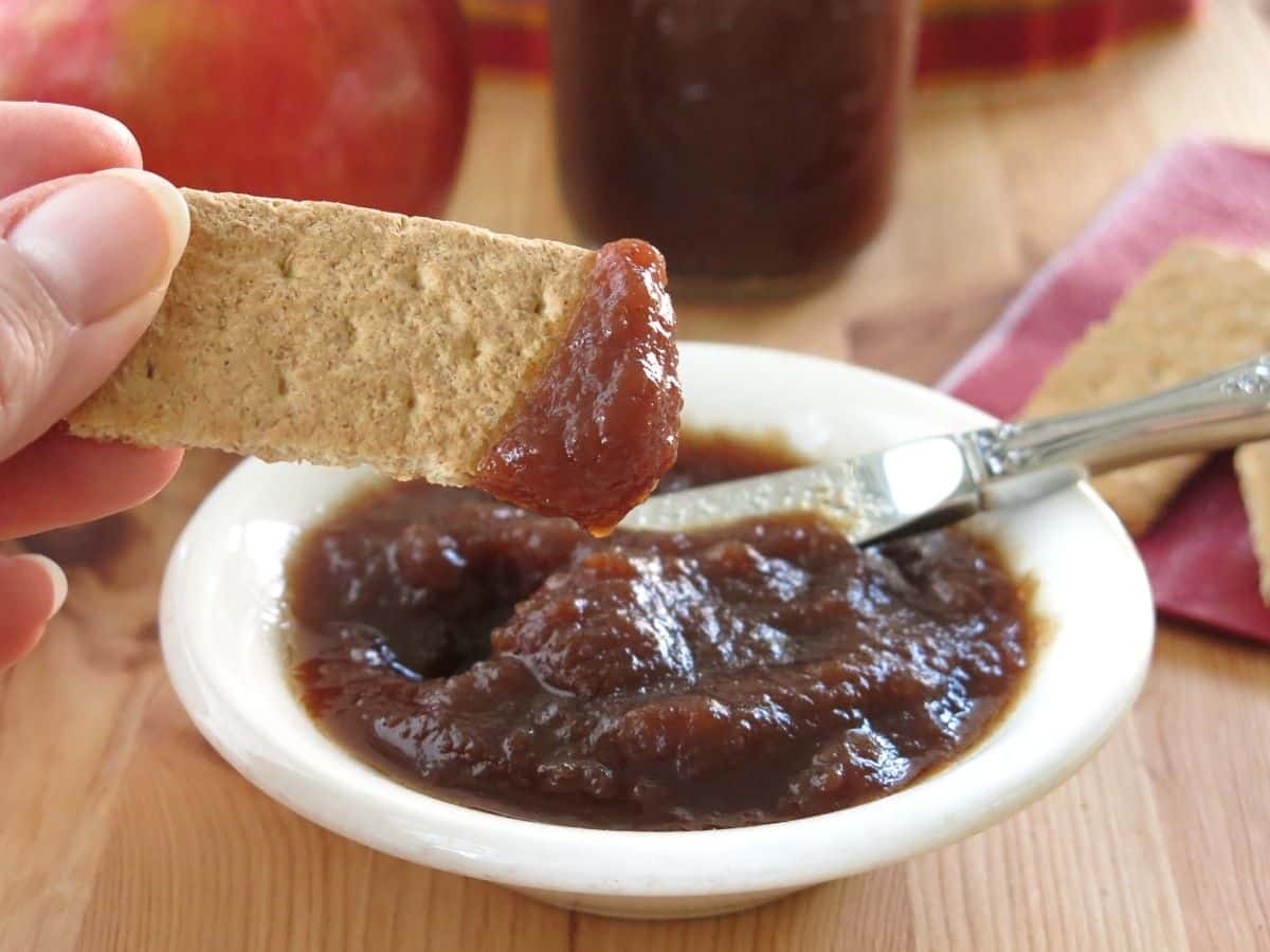 Hand holding a graham cracker dipped in low-sugar apple butter with a bowl with more apple butter below it.