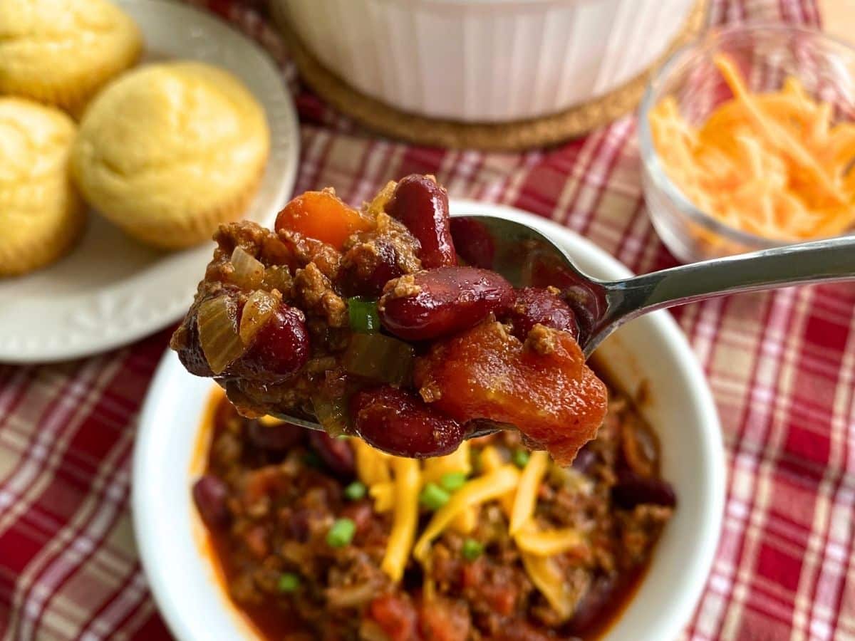 A spoon lifting easy Southern chili out of a bowl with corn muffuns and cheese behind it.
