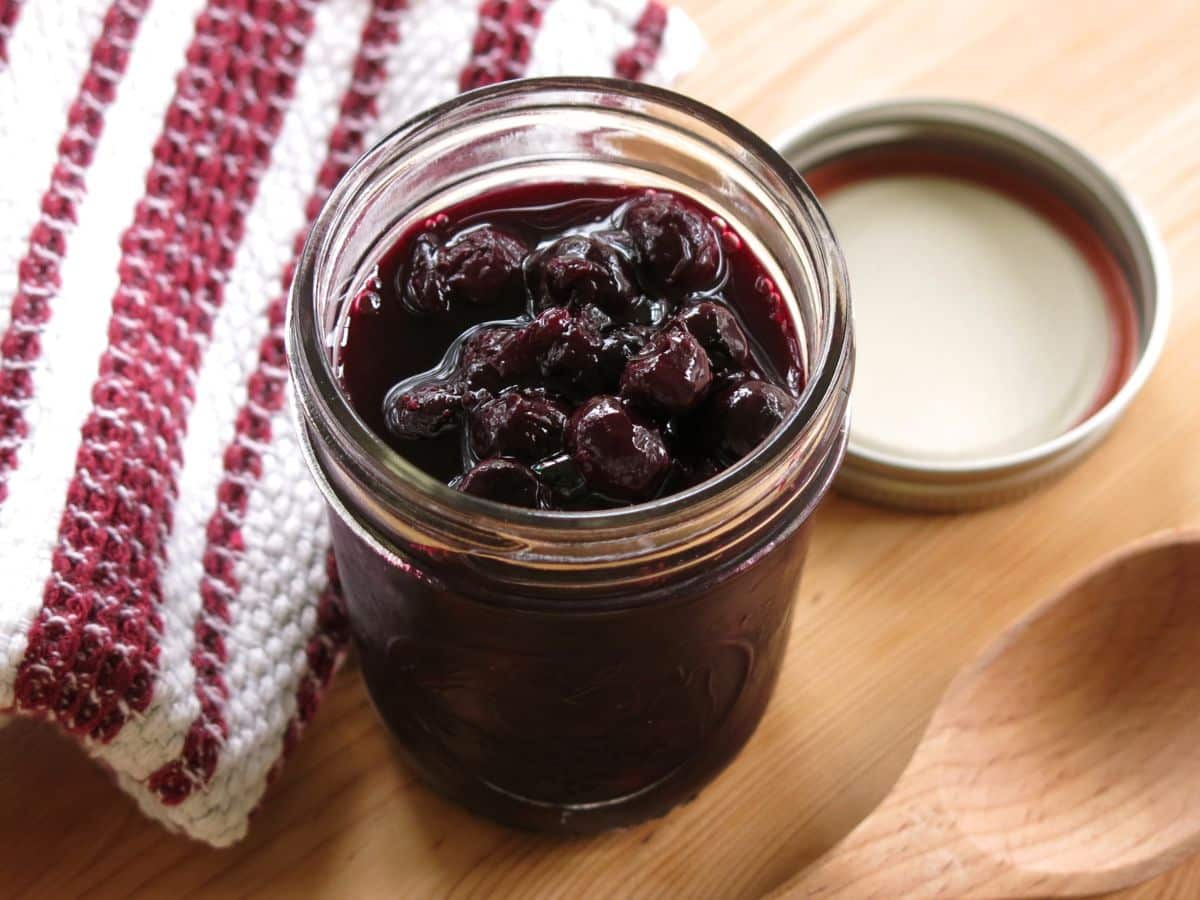 Open mason jar with blueberry compote next to the lid a rid towel and a wooden spoon.