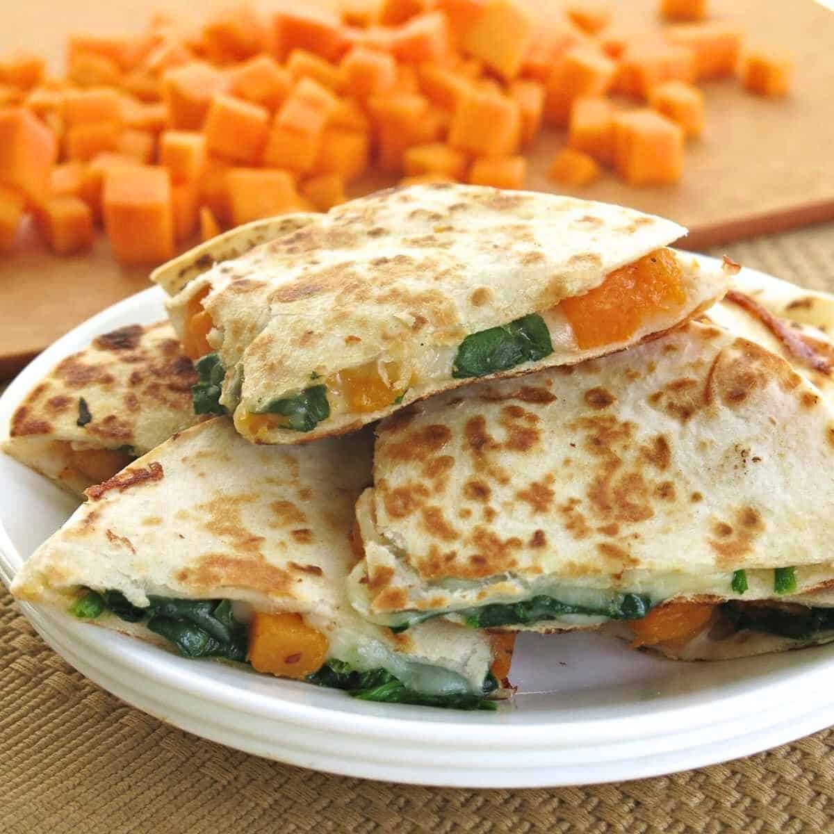 Butternut squash quesadillas piled on a plate with cubes of butternut squash in the background.