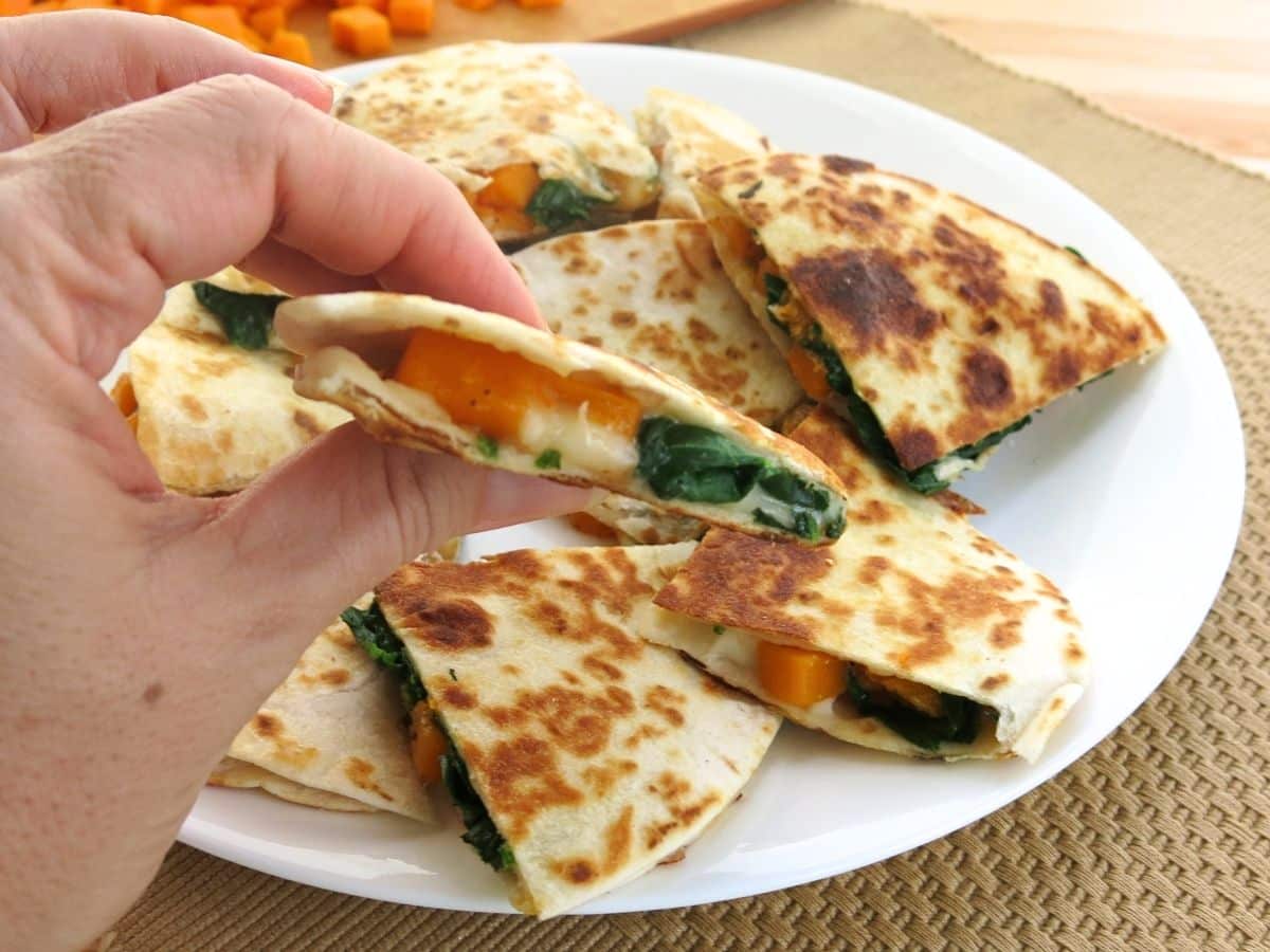 Hand holding a a triangle of a butternut squash quesadilla with spinach.