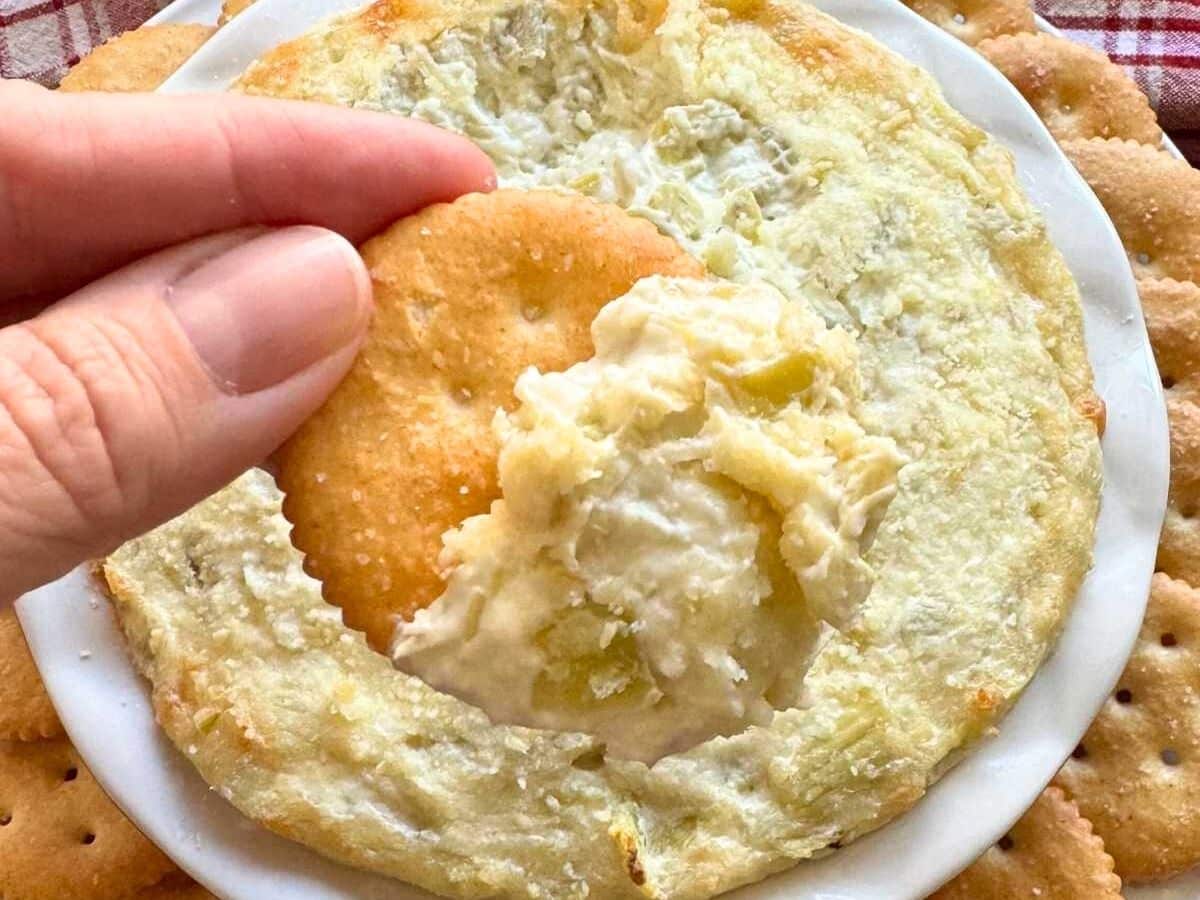 Fingers holding a cracker with artichoke dip on it over a bowl with more of it.