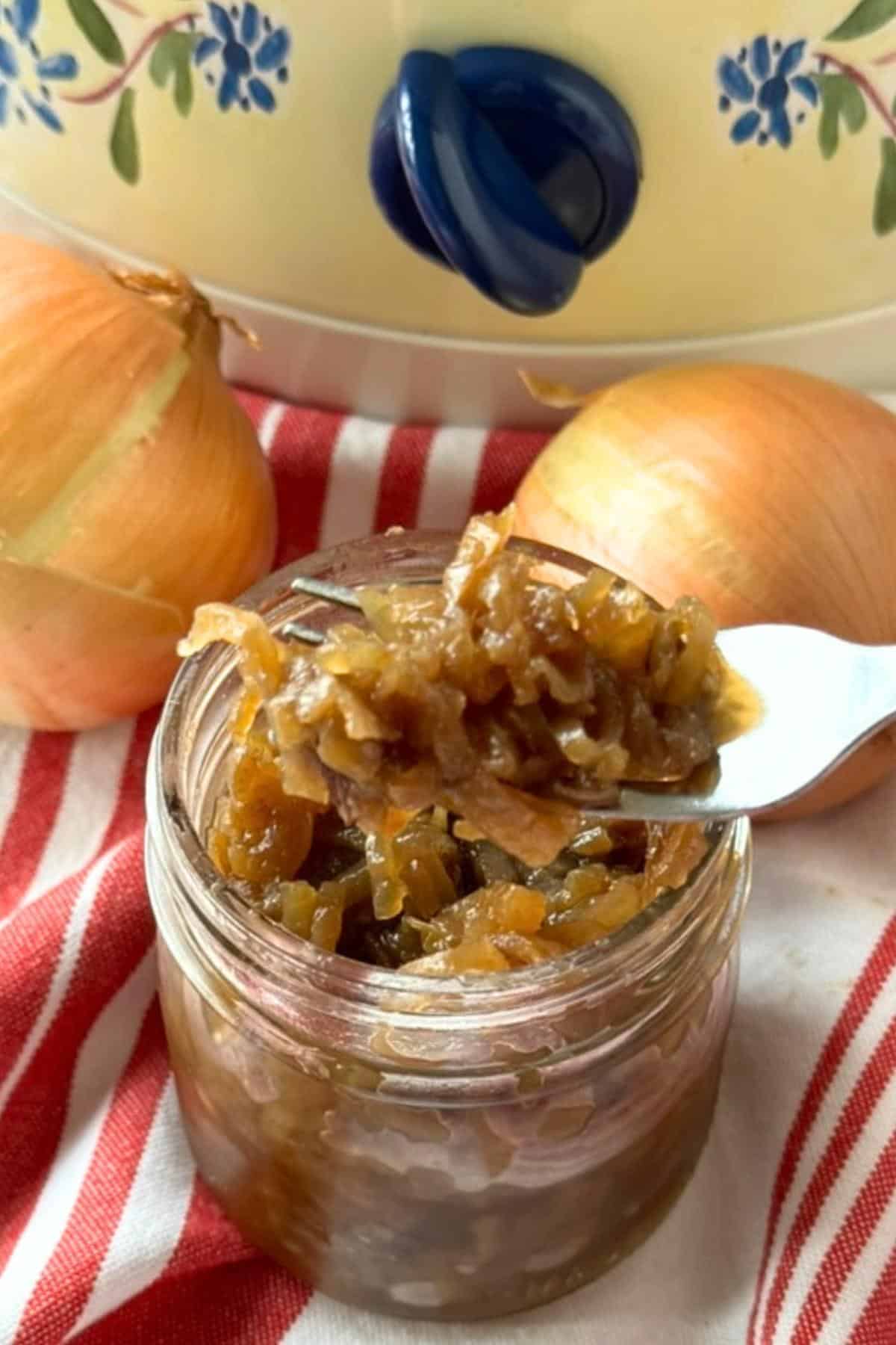 Fork lifting caramelized onions out of a glass jar with 2 yellow onions and a slow cooker behind it.