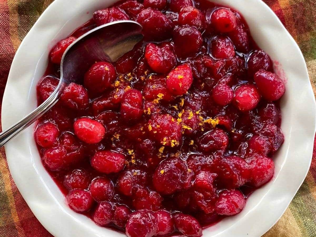 Whole cranberry sauce in a fluted bowl with zested orange and a spoon.