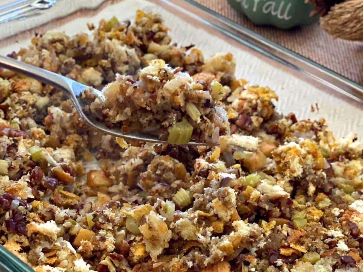 Spoon lifting stuffin with apples, celery, and pecans out of a glass baking dish.