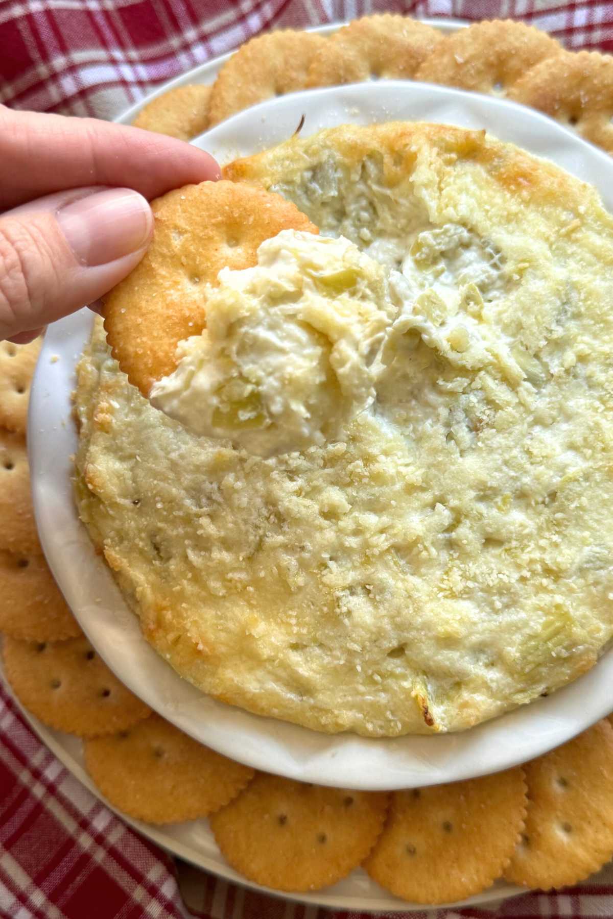 Hand holding a cracker with artichoke dip on it over the bowl of dip with Ritz crackers surrounding it.
