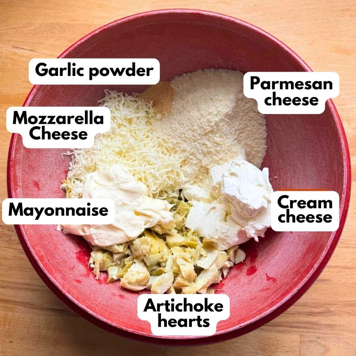 Red bowl with labeled ingredients to make artichoke dip: cream cheese, mozzarella cheese, Parmesan cheese. mayonnaise, chopped artichoke hears, and garlic powder.