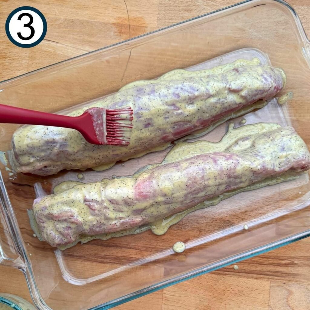 Two pork tenderloin in a glass baking dish with a red silicone brush spreading the lemon mustard sauce on top.