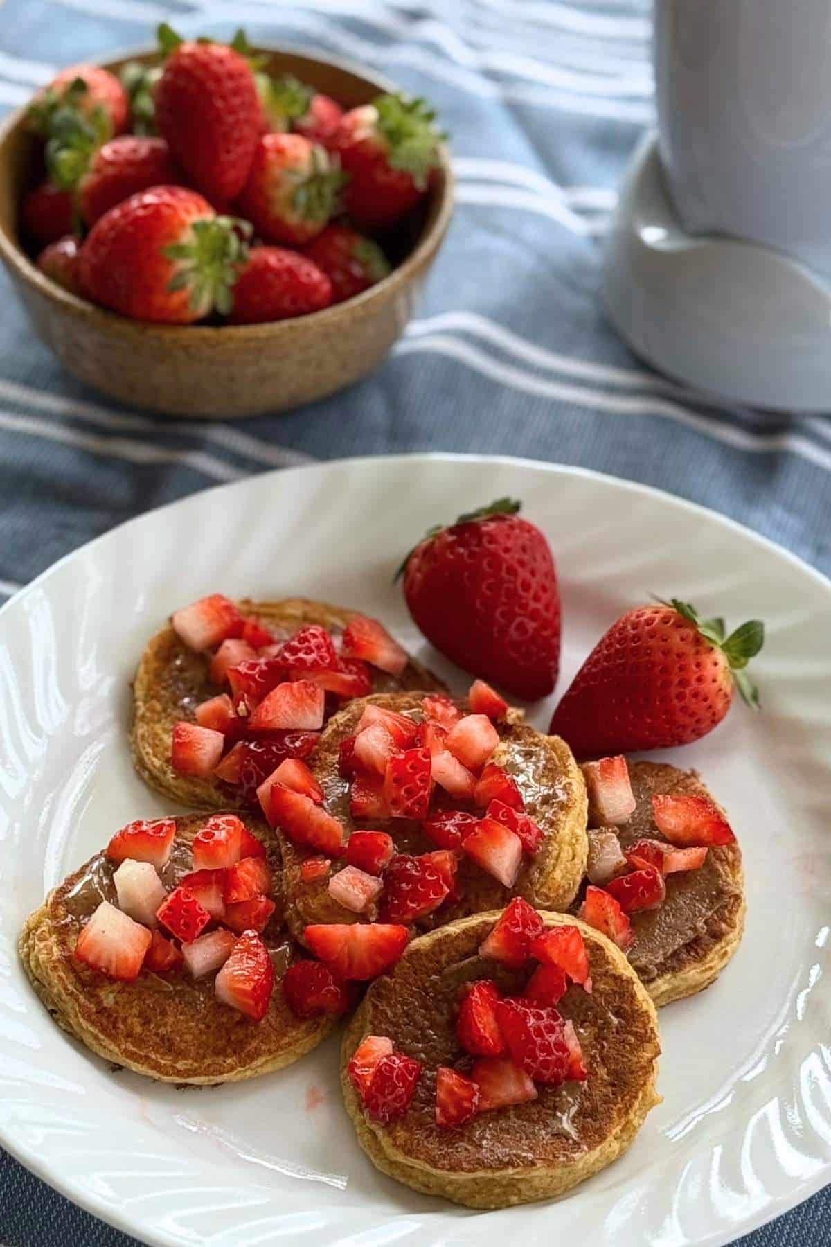 Five oat cottage cheese pancakes slathered with almond butter and topped with diced strawberries on a white plate with a blender and bowl of strawberries behind it.