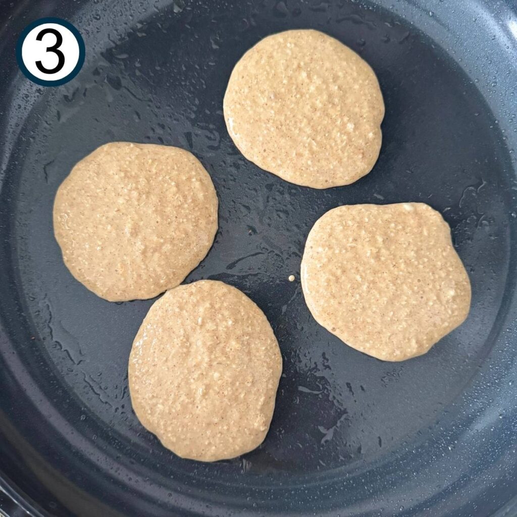 Four partially cooked cottage cheese and oat pancakes in a dark non-stick pan.
