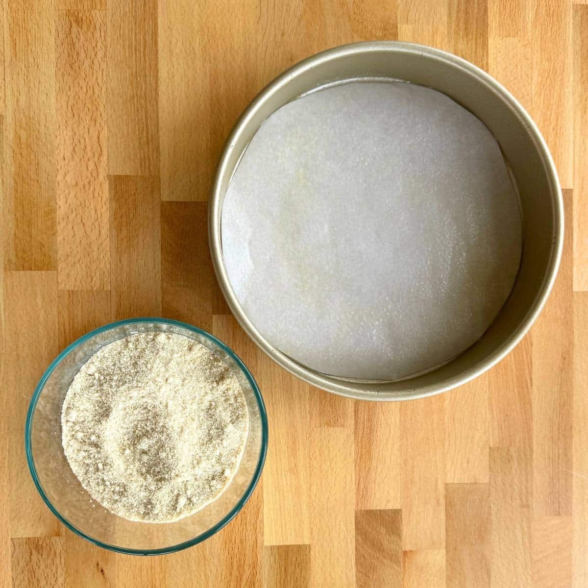 9-inch cake pan with parchment paper in the bottom and glass bowl with almond flour, baking powder, and salt.