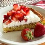 Slice of flourless lemon almond cake topped with whipped cream and strawberries on a white plate.
