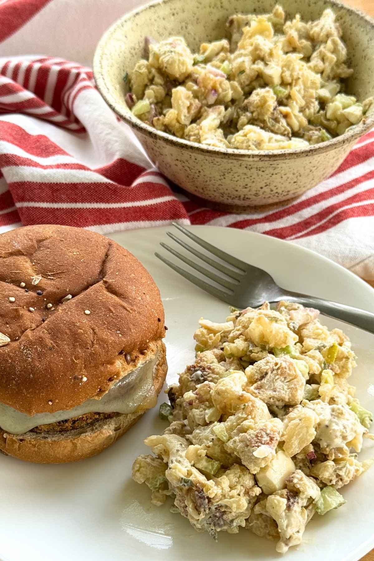 A serving of cauliflower mock potato salad on a white plate with a fork next to a hamburger and with a bowl of more cauliflower potato salad in the background.
