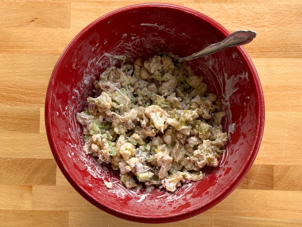 Cauliflower potato salad with dressing, relish, celery, red onion, and chopped egg in a red bowl with a large spoon.