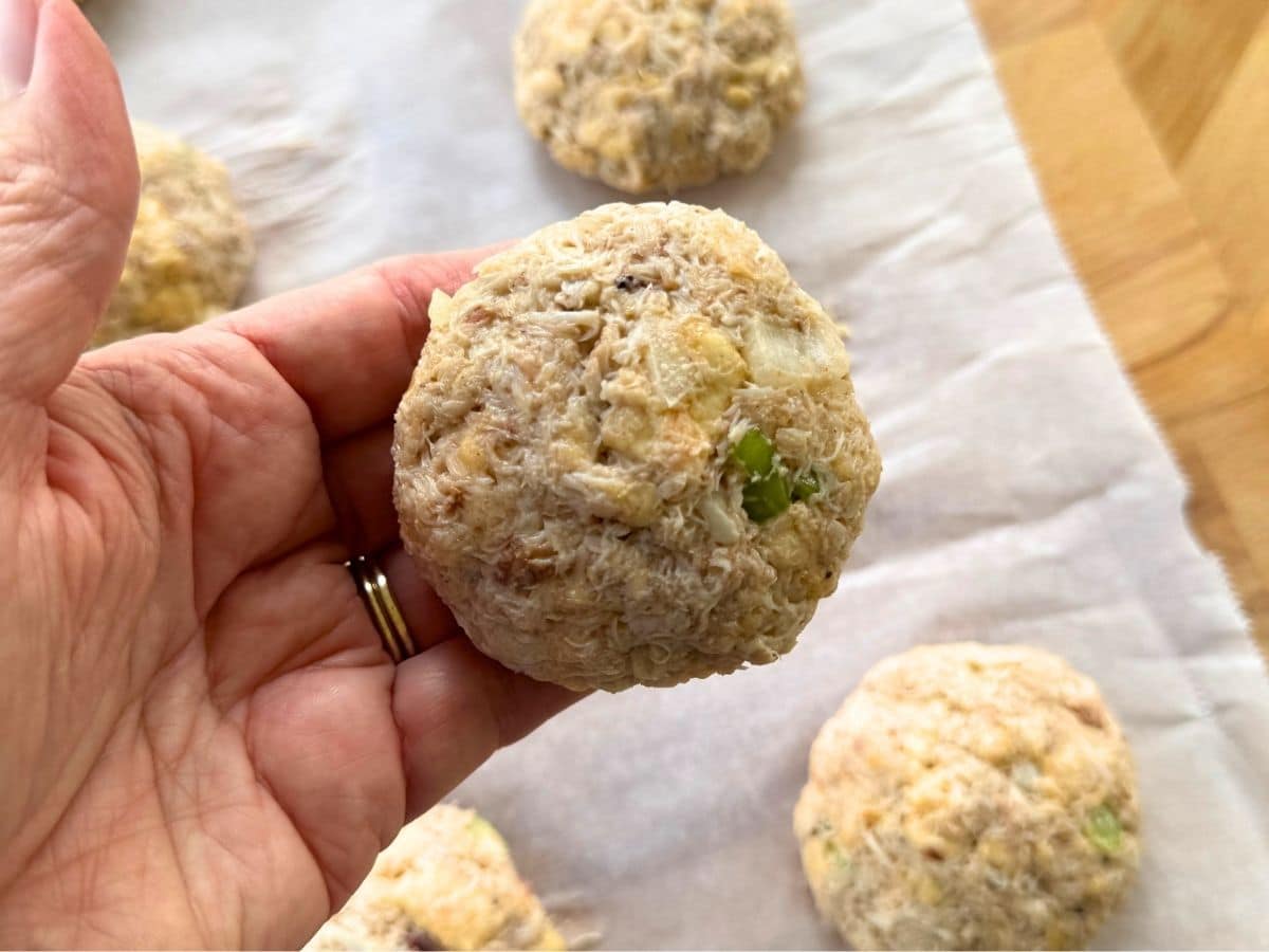 Hand holding an uncooked Maryland-style lump crab cake with a slightly domed shape over parchment paper with more crab cakes on it.
