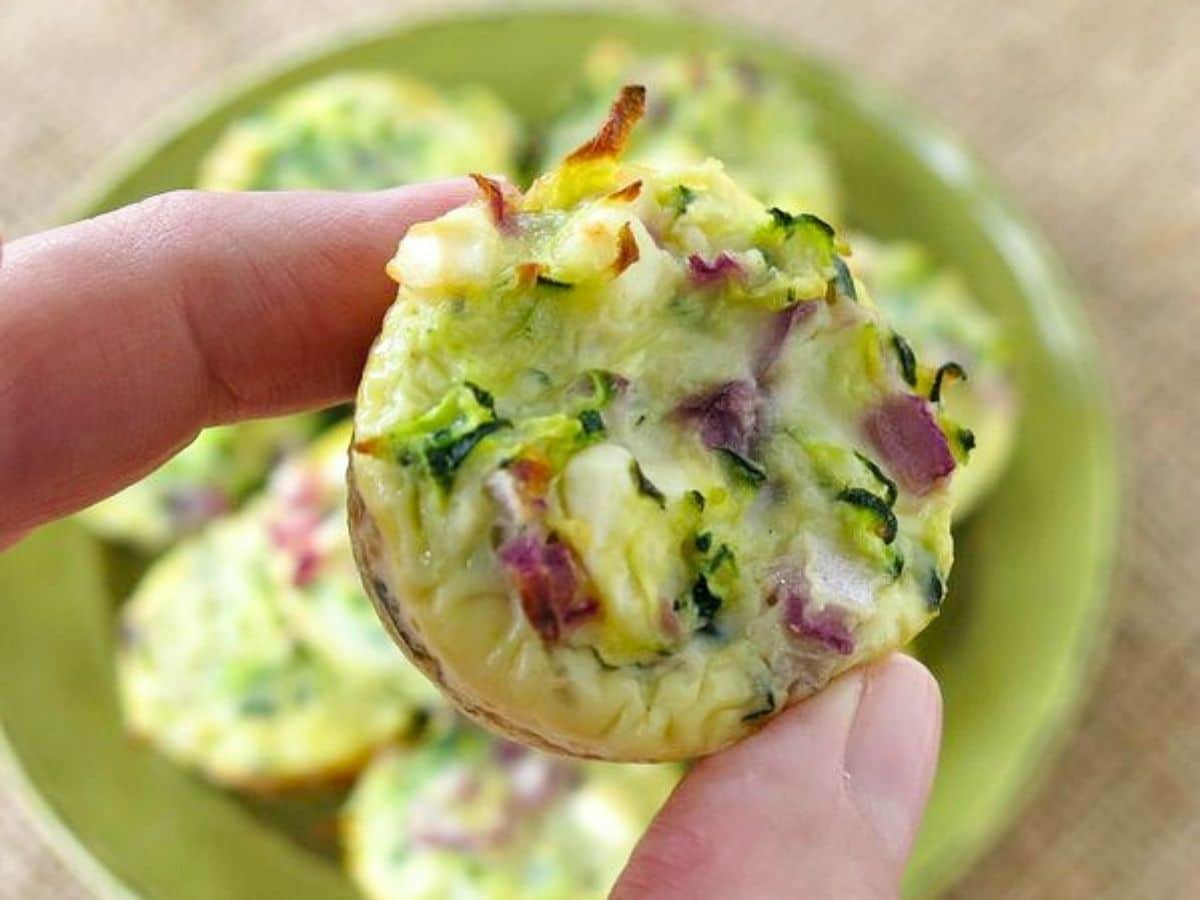 Fingers holding a zucchini egg bite over a green plate with more of them.