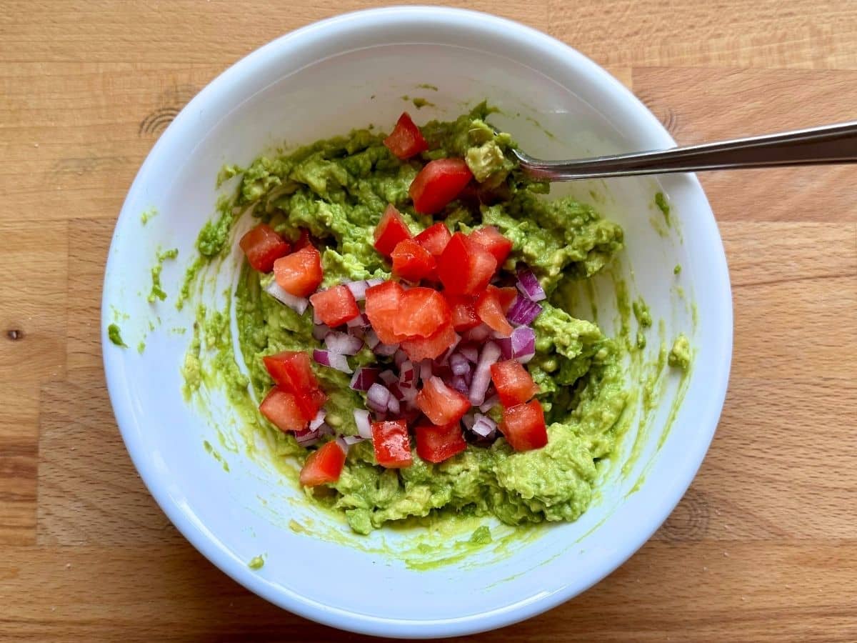 Diced red onion and diced plum tomato on top of mashed avocado in a white bowl with a fork.
