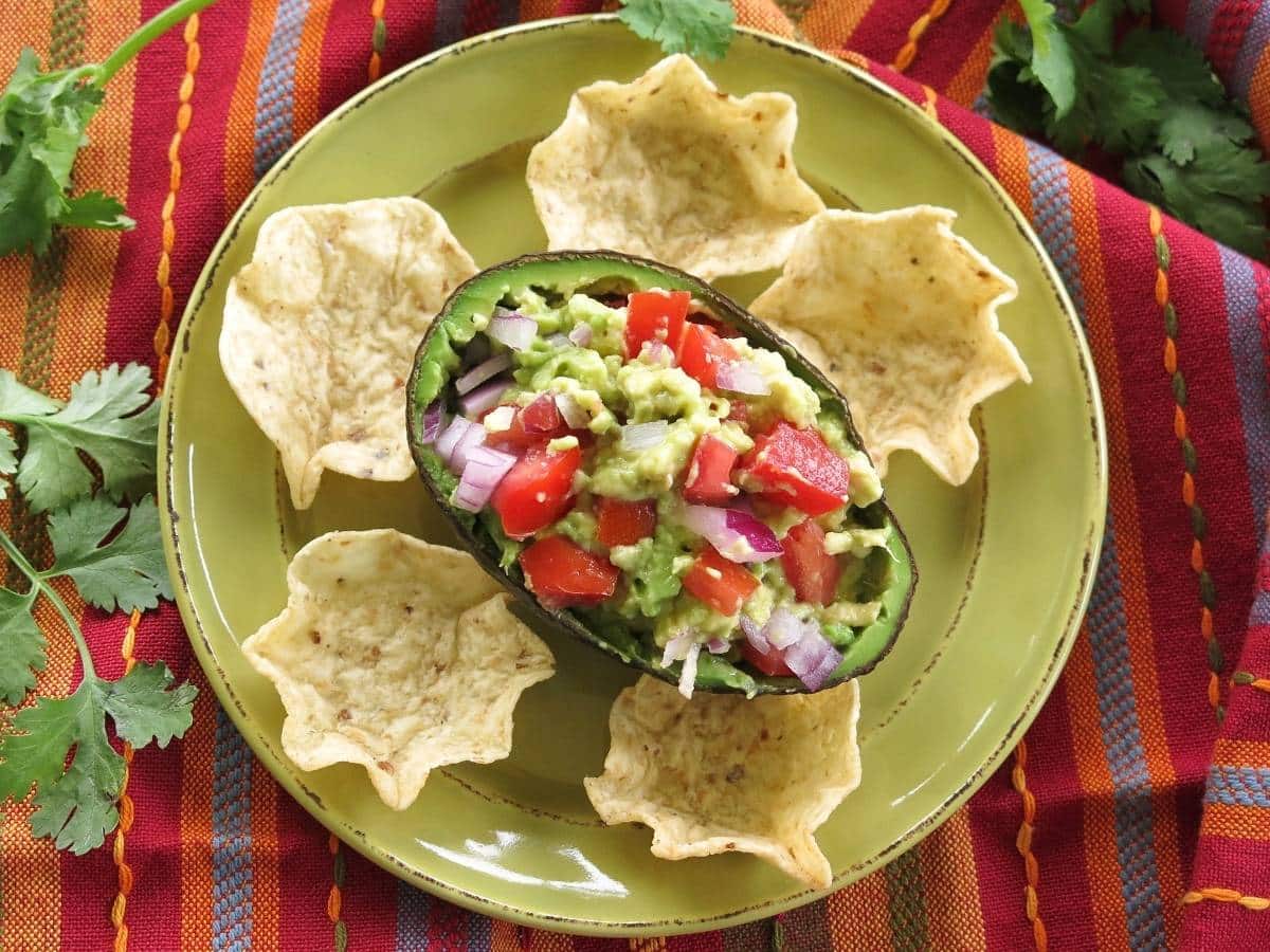 Half of a Hass avocado stuffed with guacamole on a plate with chips next to cilantro.