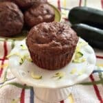 A healthy chocolate zucchini muffin made with Greek yogurt and chocolate chips on small cake pedestal surrounding by shreds with zucchini and more muffins in the background.