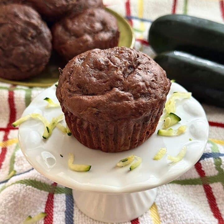 A healthy chocolate zucchini muffin made with Greek yogurt and chocolate chips on small cake pedestal surrounding by shreds with zucchini and more muffins in the background.