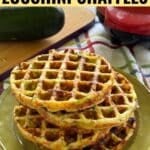 Four zucchini chaffles stacked on a green plate with a Dash waffle maker and a zucchini in the background.