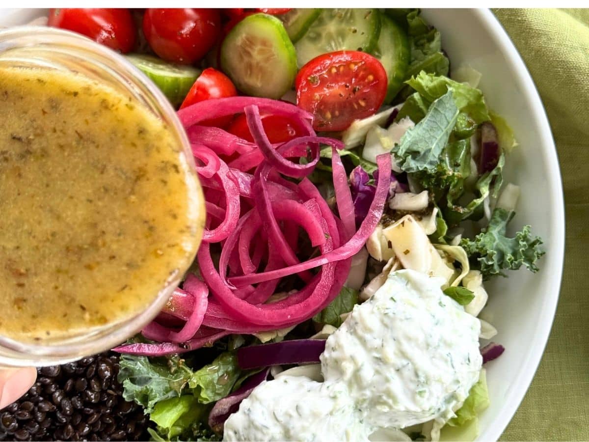 Greek vinaigrette being poured over assembled Mediterranean bowl.