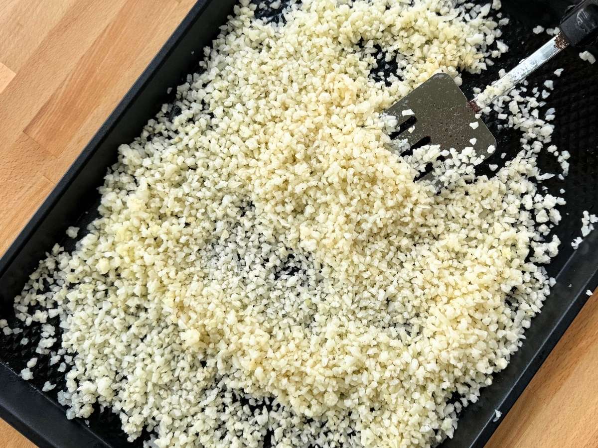 Partially cooked cauiflower rice being lifted with a spatula over a large sheet pan.