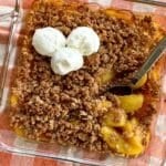 Peach crisp with an almond flour and oat topping in a glass dish with a spoon lifting peaches that were cooked from frozen.