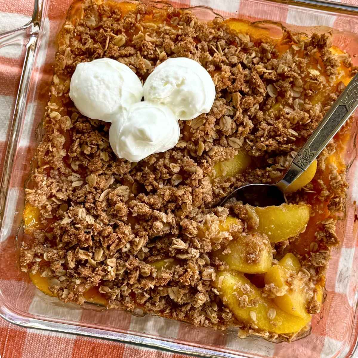 Peach crisp with an almond flour and oat topping in a glass dish with a spoon lifting peaches that were cooked from frozen.