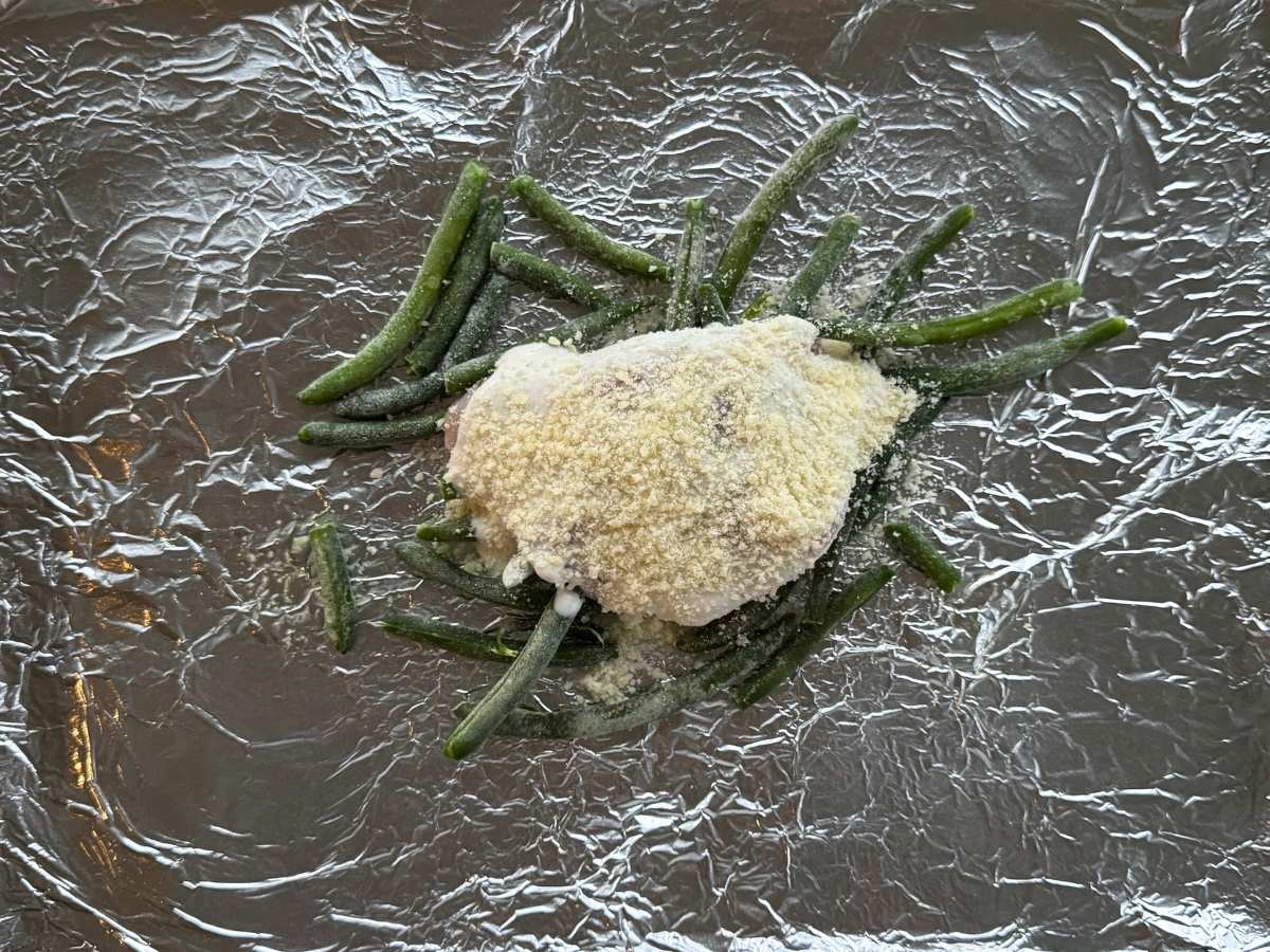 Uncooked chicken topped with ranch dressing and Parmesan cheese on top of green beans on heavy aluminum foil.