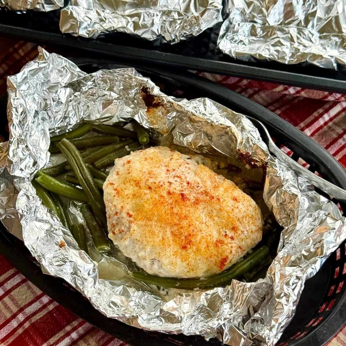 Ranch chicken foil packet opened to expose chicken topped with ranch dressing and Parmesan cheese over green beans with more packets in the background.