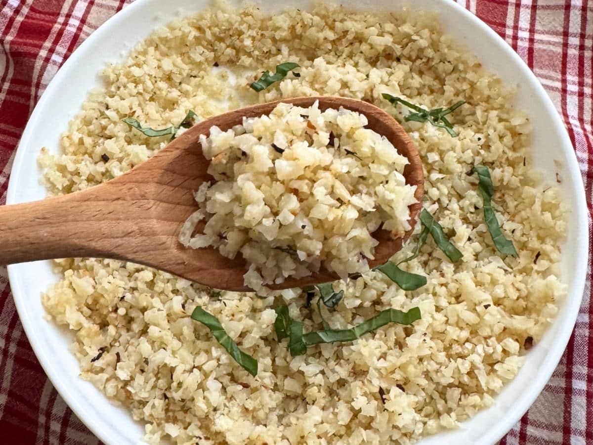 Wooden spoon lifting roasted riced cauliflower from a white bowl.