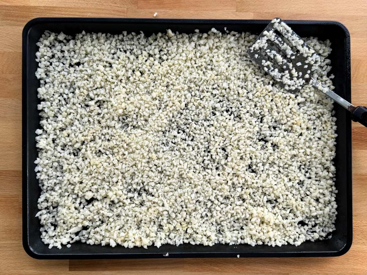 Frozen cauliflower coated with olive oil and sprinkled with salt, pepper, and garlic powder on a large sheet pan with a spatula.