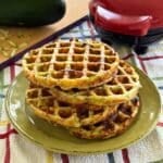 Four zucchini chaffles stacked on a green plate with a Dash waffle maker and a zucchini in the background.