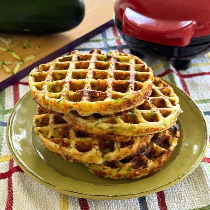 Four zucchini chaffles stacked on a green plate with a Dash waffle maker and a zucchini in the background.