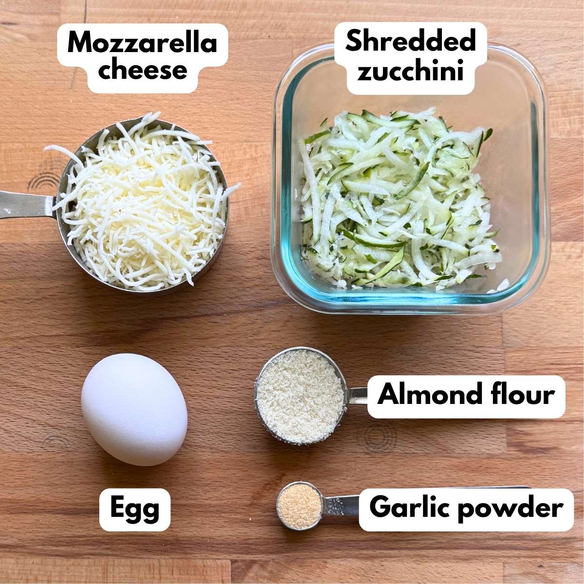 Shredded zucchini, shredded mozzarella cheese, egg, almond flour, and garlic powder to make zucchini chaffles on a cutting board.
