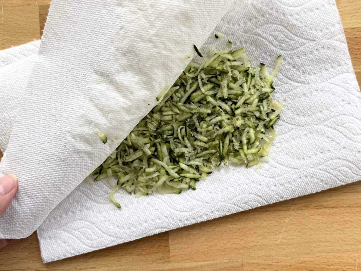 Shredded zucchini between two paper towels with the top paper towel being lifted up.
