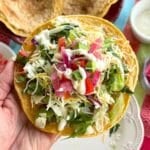 Hand holding a crispy ground chicken taco topped with lettuce, cheese, lime crema, red onions, and tomatoes.