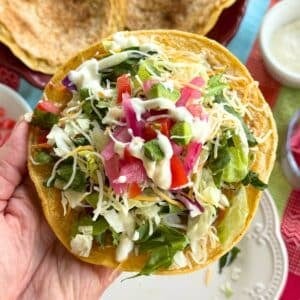 Hand holding a crispy ground chicken taco topped with lettuce, cheese, lime crema, red onions, and tomatoes.