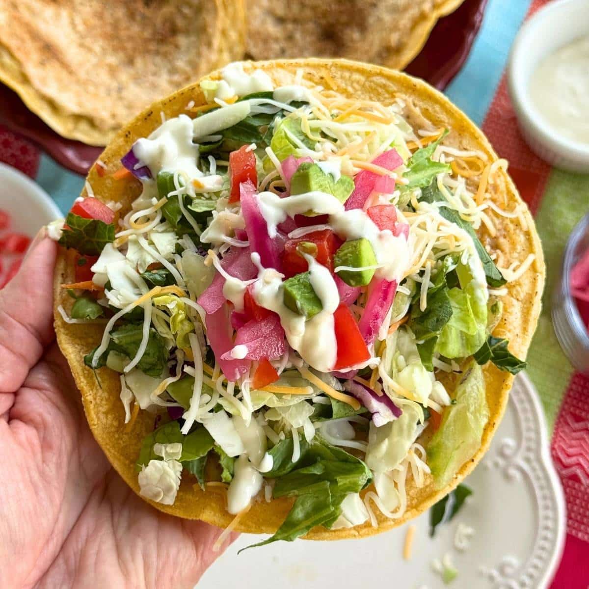 Hand holding a crispy ground chicken taco topped with lettuce, cheese, lime crema, red onions, and tomatoes.