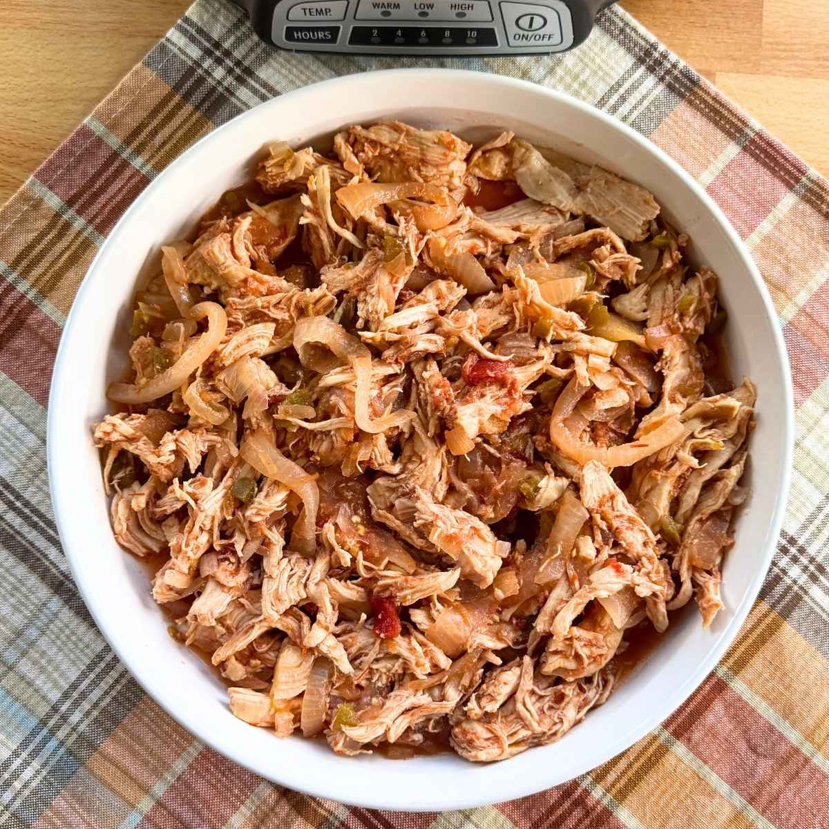 Shredded slow cooker salsa chicken with cooked onions in a serving bowl in front of the control panel for a a slow cooker.