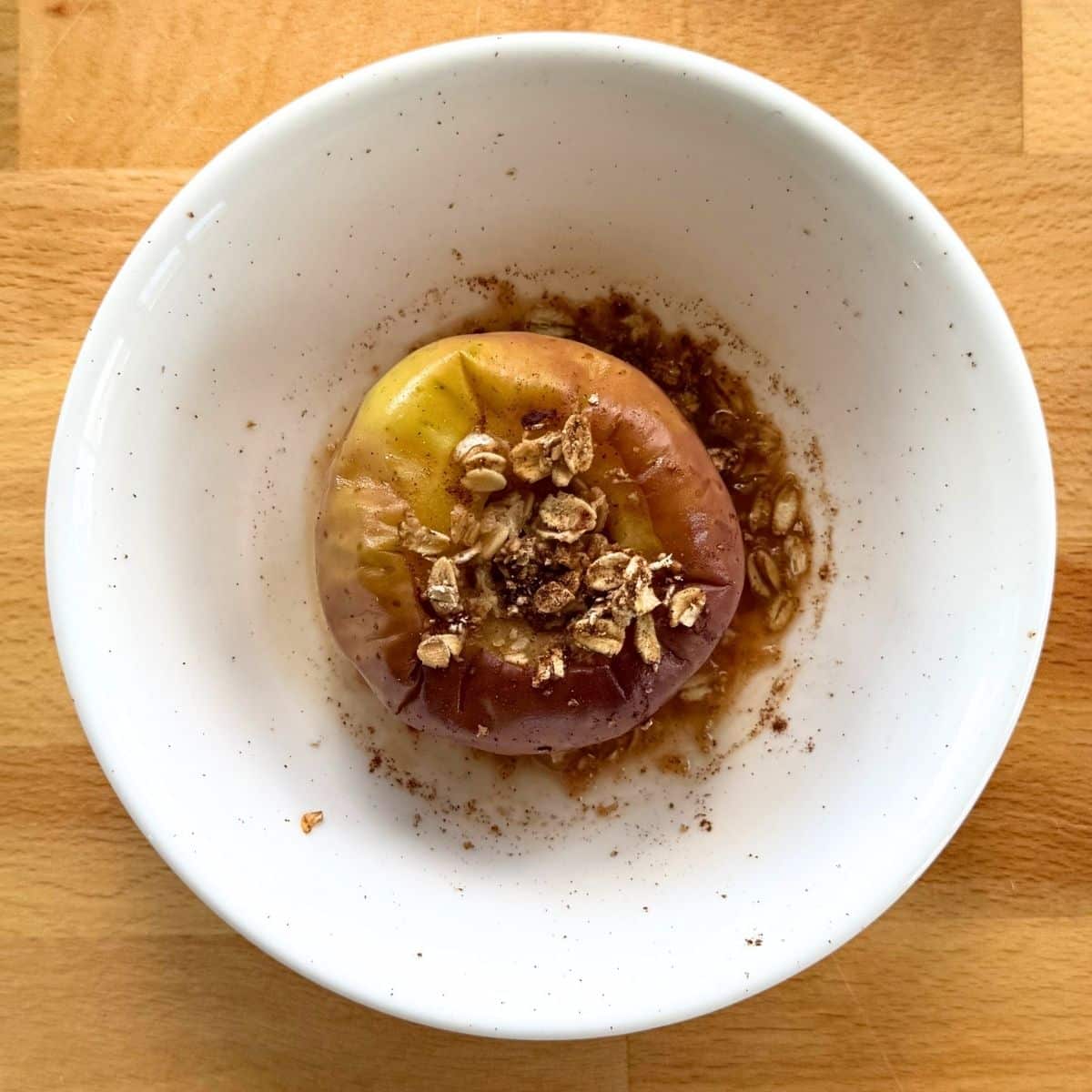 Microwave baked apple stuffed with oats and cinnamon in a white bowl exposing a little juice under it after cooking.