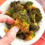 Hand holding a crispy Parmesan broccoli bite with golden, cheesy edges over a bowl of roasted broccoli.